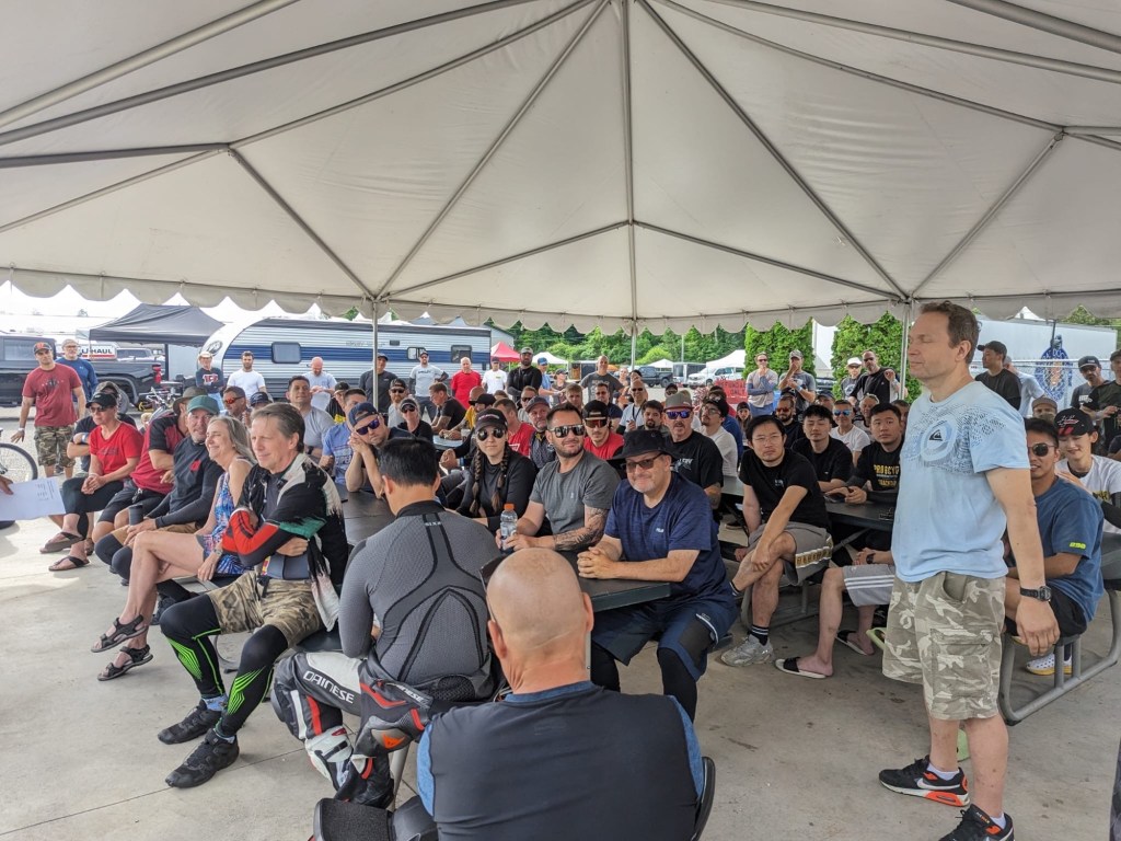 Track day paddock scene with riders gathered, bikes parked, community atmosphere